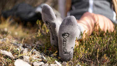 Une personne est allongée sur le dos dans l'herbe, les pieds tendus vers l'avant, chaussés de chaussettes de randonnée grises avec un logo noir et l'inscription « GIESSWEIN ». À l'arrière-plan, on aperçoit floues des vêtements de randonnée et la nature.