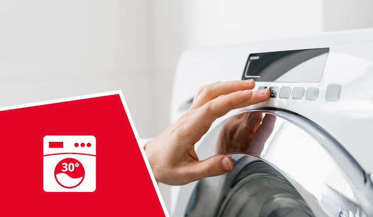 Une main réglant le bouton de commande d'une machine à laver blanche. Une icône rouge représente le symbole d'une machine à laver avec la mention « 30° », indiquant que le lavage en machine est possible à 30 °C.
