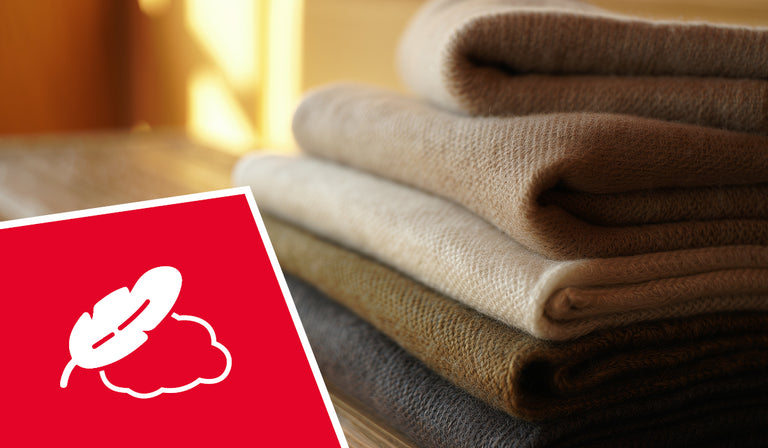 Une pile de pulls ou de vêtements en laine tricotée beige et marron, soigneusement pliés, repose sur une surface en bois baignée d'une lumière naturelle et chaleureuse. Au premier plan, un icône carré rouge représente un symbole blanc en forme de feuille barrée d'un nuage, indiquant des propriétés hypoallergéniques ou sans allergènes. Les tissus doux et texturés semblent confortables et luxueux dans la lumière dorée ambiante.