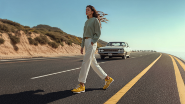 Femme en pull gris et pantalon blanc traversant la rue avec des baskets jaunes, voiture vintage en arrière-plan