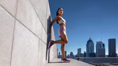 Femme en tenue de sport appuyée contre un mur en béton sur un toit, portant des baskets Wool Peak bordeaux, avec la silhouette d'une grande ville en arrière-plan.