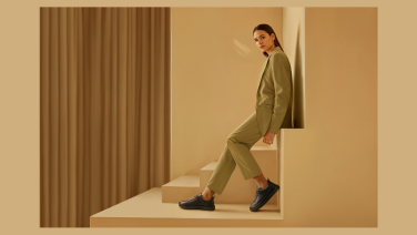 Une jeune femme vêtue d'un tailleur vert olive est assise sur un podium beige dans une pièce au design minimaliste. Elle porte des chaussures noires. La scène dégage une impression de modernité et d'élégance avec ses formes géométriques épurées.