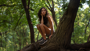 Une jeune femme aux longs cheveux foncés est assise, souriante, sur une grosse branche d'arbre dans la forêt. Elle porte des baskets foncées.