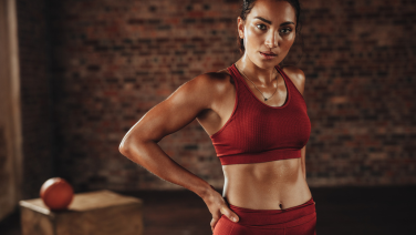 Une femme sportive vêtue d'une tenue de sport rouge se tient debout avec assurance, la main sur la hanche, dans une salle de sport devant un mur en briques.