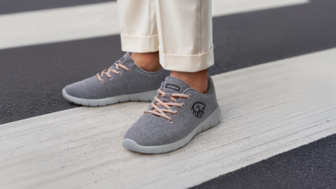 Des pieds chaussés de baskets Giesswein grises à lacets orange se tiennent sur un passage piéton blanc. La personne porte un pantalon blanc et les chaussures arborent le logo caractéristique de Giesswein.