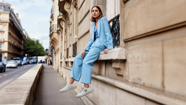 Une femme vêtue d'un tailleur bleu clair est assise, détendue, sur une balustrade en pierre devant une architecture classique. Elle porte des baskets blanches et sourit à la caméra. En arrière-plan, on aperçoit une élégante rue urbaine.