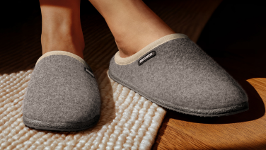 Gros plan sur des pieds chaussés de pantoufles Giesswein grises posés sur un tapis beige texturé. Les pantoufles ont une semelle foncée et arborent le logo caractéristique de Giesswein.