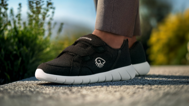 Gros plan sur des pieds chaussés de baskets Giesswein noires à semelles blanches sur un chemin de gravier. La personne porte un pantalon marron et, dans l'arrière-plan flou, on aperçoit des plantes vertes et des montagnes.