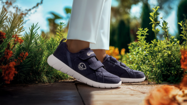 Gros plan sur des pieds chaussés de baskets Giesswein bleu foncé à semelles blanches, posés sur une terrasse en bois. La personne porte un pantalon blanc. Plantes vertes floues et fleurs orange en arrière-plan.
