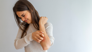Une femme aux longs cheveux bruns, vêtue d'un pull-over beige en tricot, se gratte l'avant-bras. Elle regarde la zone qui la démange tout en se grattant le bras de l'autre main. Fond gris neutre.