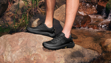 Deux pieds chaussés de chaussures de randonnée noires (Wool Trail X de Giesswein) se tiennent sur un rocher brun au bord d'un petit ruisseau où coule de l'eau.