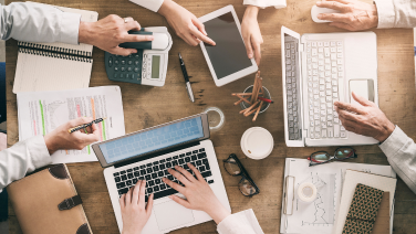 Plusieurs personnes travaillent ensemble à une table avec des ordinateurs portables, une tablette, une calculatrice et des documents - une atmosphère d'équipe très animée.