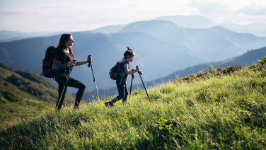 Deux randonneuses équipées de sacs à dos et de bâtons de randonnée gravissent un versant herbeux. À l'arrière-plan, un vaste paysage montagneux composé de plusieurs chaînes de montagnes aux teintes bleutées s'étend sous un ciel nuageux.