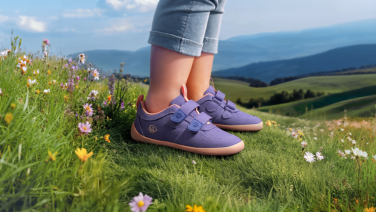 Chaussures pour enfants violettes avec fermeture velcro sur une prairie verte parsemée de fleurs sauvages colorées. À l'arrière-plan, un paysage vallonné s'étend sous un ciel bleu, avec de douces collines verdoyantes.