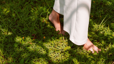 Une personne en pantalon blanc marche dans une végétation luxuriante et touche de jeunes plantes avec ses deux mains. La scène montre un jardin naturel avec différentes plantes vertes, herbes et plantes sauvages dans différentes nuances de vert.