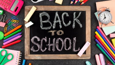 Une ardoise noire avec l'inscription « BACK TO SCHOOL » écrite à la craie blanche est posée au milieu d'un fond sombre. Divers accessoires scolaires sont disposés tout autour.