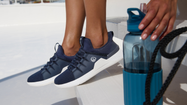 Gros plan sur des pieds chaussés de baskets bleu foncé à côté d'une bouteille d'eau turquoise sur un fond clair. Une main attrape la bouteille.