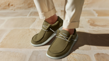 Gros plan sur des pieds chaussés de pantalons blancs et de chaussures vert olive à lacets blancs et semelles claires. La personne se tient debout sur un sol en pierre claire.