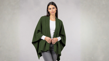 Femme portant une cape Giesswein Merino vert foncé sur un haut blanc et un pantalon gris devant un fond gris clair.