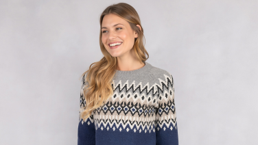 Femme souriante aux longs cheveux blonds portant un pull Giesswein gris avec un motif norvégien bleu marine sur fond clair