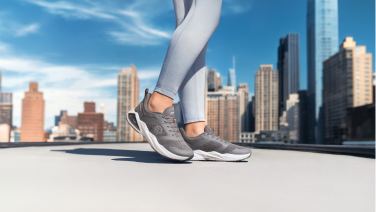 Gros plan sur des baskets grises Giesswein Merino Wool aux pieds d'une coureuse vêtue d'un legging bleu clair, sur une terrasse sur le toit avec des gratte-ciel en arrière-plan.