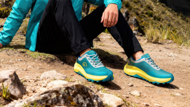 Une personne est assise sur un rocher dans les montagnes et porte des baskets Giesswein turquoise et jaunes, un pantalon noir et une veste turquoise.