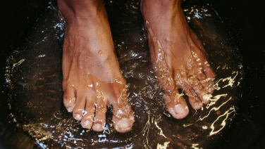 Les pieds nus d'une personne debout dans une eau claire et éclaboussante. Les gouttes d'eau et les éclaboussures sont figées dans leur mouvement, ce qui souligne l'atmosphère rafraîchissante et naturelle.
