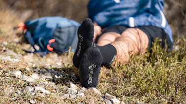 Une personne est allongée, détendue, sur un sol rocheux en pleine nature. Au premier plan, on aperçoit ses pieds chaussés de chaussettes en laine mérinos noires de la marque Giesswein.
