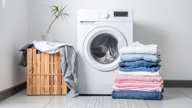 Une machine à laver blanche dans une salle de bains lumineuse, entourée de linge plié, d’une caisse en bois et d’une plante verte.