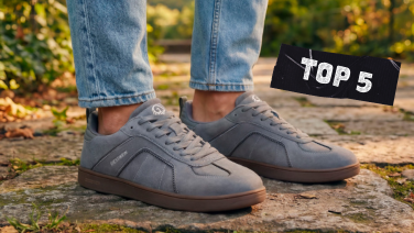 Gros plan sur les deux pieds chaussés de baskets en daim gris clair, dotées d'une semelle en caoutchouc marron et du logo Giesswein blanc, sur des pavés baignés d'une lumière automnale.