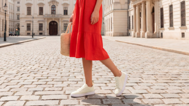 Une femme vêtue d'une robe d'été rouge et portant un sac en osier traverse une ruelle pavée de la vieille ville, chaussée de baskets en tricot blanc.