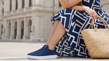 Une femme vêtue d'une robe d'été à motifs bleus et blancs est assise sur une marche devant un bâtiment historique de la ville ; elle porte des baskets en tricot bleu marine et tient à la main la poignée d'un sac en osier tressé.