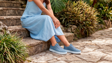 Gros plan sur des pieds chaussés de baskets en tricot bleu clair à semelle blanche, posés sur des marches en pierre, avec en arrière-plan une robe en lin bleu clair et un chemin pavé.
