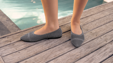 Des pieds chaussés de ballerines en tricot gris, posés sur une passerelle en bois au bord d'une piscine.