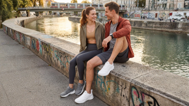 Un couple est assis sur un muret au bord d'une rivière en ville, tous deux rient ; ils portent tous les deux des baskets grises Giesswein Merino Runner.