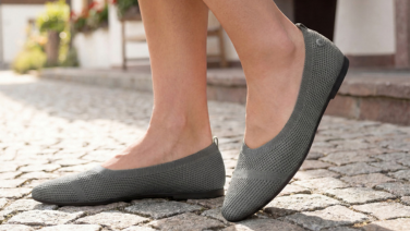 Gros plan sur des pieds chaussés de ballerines en maille grise de la marque Giesswein, posés sur un pavé.
