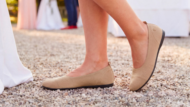 Gros plan sur des pieds chaussés de ballerines en maille beiges de la marque Giesswein, marchant sur un chemin de galets ; à l'arrière-plan, une table dressée pour une fête.