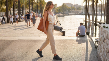 Une femme, munie d'un sac et d'un tapis de yoga, se promène le long d'une promenade en bord de mer bordée de palmiers ; elle porte des baskets noires Giesswein Wooltastic.