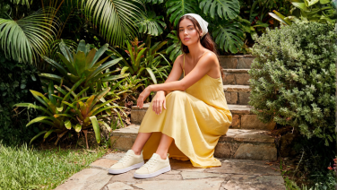 Une femme est assise sur un escalier en pierre dans un jardin luxuriant. Elle porte une robe d'été jaune, un bandeau blanc et des baskets Giesswein couleur crème.