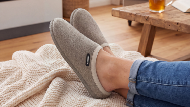 Gros plan sur les pieds croisés d'une personne en jean, allongée sur une couverture en tricot de couleur crème ; à l'arrière-plan, une table en bois avec une boisson et un livre. Elle porte des pantoufles en laine mérinos grise bordées d'une bande claire.