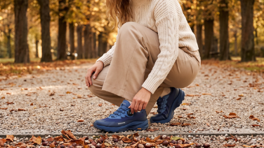 Une personne ajuste sa chaussure de randonnée bleue à semelle Vibram sur un chemin de gravier parsemé de feuilles et de châtaignes.