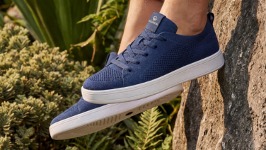 Gros plan sur une paire de baskets Giesswein en laine mérinos bleu foncé, dotées d'une semelle plate blanche, devant un décor de végétation méditerranéenne et de pierres.