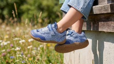 Gros plan sur une basket enfant Giesswein bleu clair avec fermeture velcro et semelle couleur miel, portée avec un jean devant une prairie estivale en fleurs.