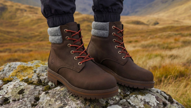 Gros plan sur les pieds d'une personne debout sur un rocher, dans un paysage de montagne automnal. Elle porte des bottines en cuir marron avec des lacets rouges, un col en feutre de laine gris et une semelle profilée.