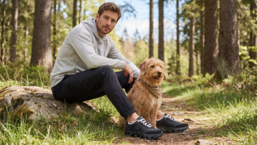 Un homme vêtu d'un sweat à capuche gris et d'un pantalon foncé est assis sur un tronc d'arbre dans la forêt, à côté d'un Goldendoodle, et porte des baskets de plein air Giesswein noires.
