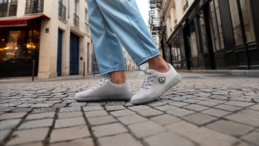 Gros plan sur des pieds chaussés de baskets Giesswein Merino Runners grises sur des pavés dans une rue urbaine.