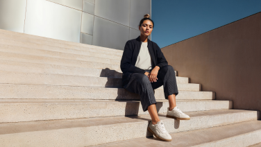 Une femme vêtue d'un blazer bleu foncé et d'un pantalon gris est assise sur des marches en pierre devant un bâtiment moderne ; elle porte des baskets blanches Giesswein en laine mérinos.
