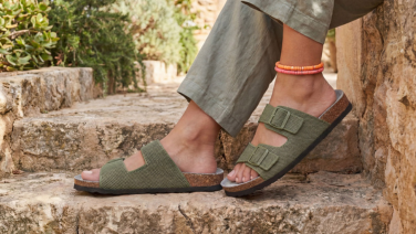 Des pieds chaussés de sandales en tricot vert olive à deux boucles et semelle en liège, posés sur des marches en pierre méditerranéennes, accompagnés d'un pantalon en lin vert olive et de bracelets de cheville colorés.