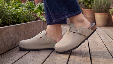 Gros plan sur les pieds d'une personne vêtue d'un jean bleu foncé, debout sur une terrasse en bois. Elle porte des sabots en laine mérinos de couleur beige, dotés de lanières à boucles et d'une semelle en liège.