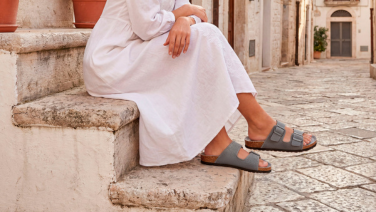Une femme en robe blanche est assise sur un escalier en pierre dans une ruelle méditerranéenne ; elle porte des sandales Giesswein grises à deux lanières et semelle en liège.